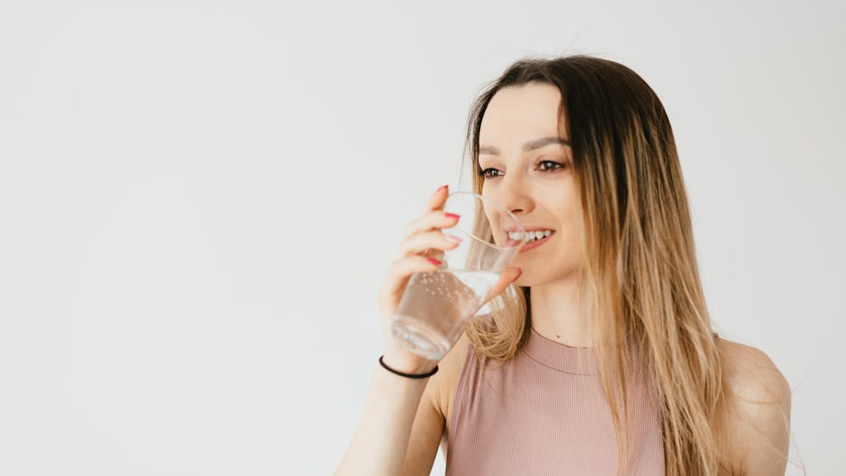 Optimistic young lady in top smiling while drinking glass of sparkling water and looking away