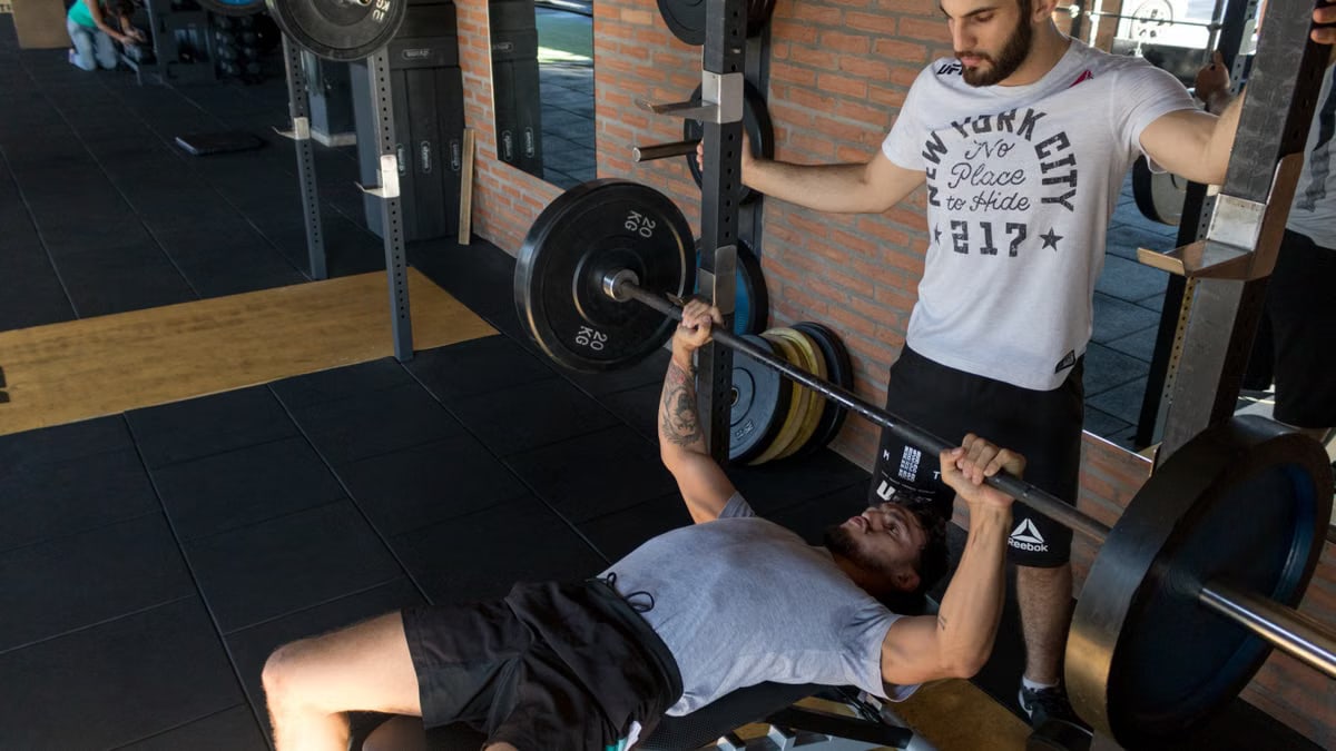 A strong man lifts weights on a bench while a trainer assists in a modern gym setup.